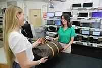 A customer hands a Louis Vuitton Monogram canvas duffle bag to a smiling store associate across a counter, with rows of laptops and electronics displayed on shelves in the background