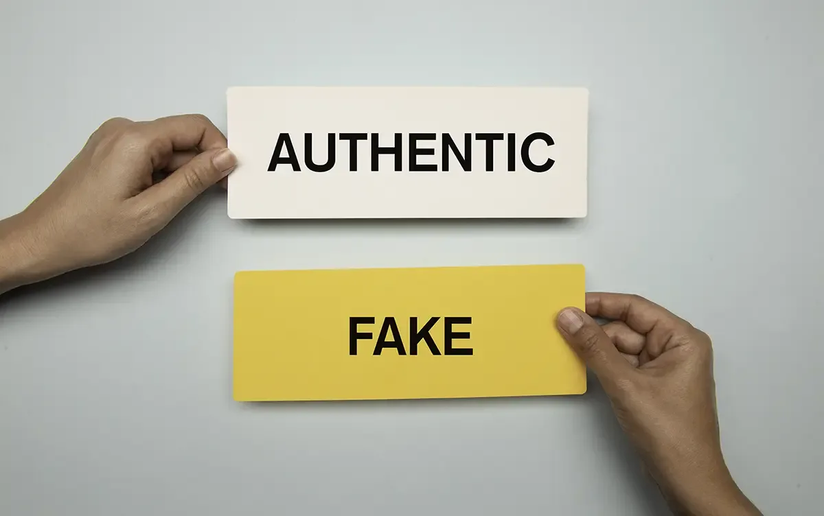Two hands holding rectangular cards labeled "AUTHENTIC" (white) and "FAKE" (yellow) side by side against a gray background, representing the concept of authenticity versus counterfeit.