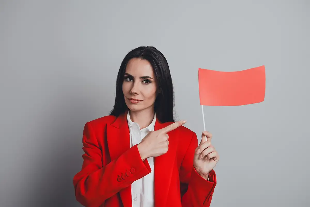 A woman in a red blazer points to a small red flag she's holding against a gray background, symbolizing a warning or red flag alert.