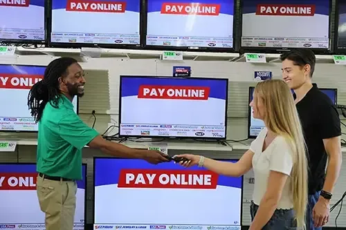 Store associate in green polo helps two customers in the electronics section, handing over a TV remote in front of a wall of flat-screen TVs displaying "Pay Online" on screen.
