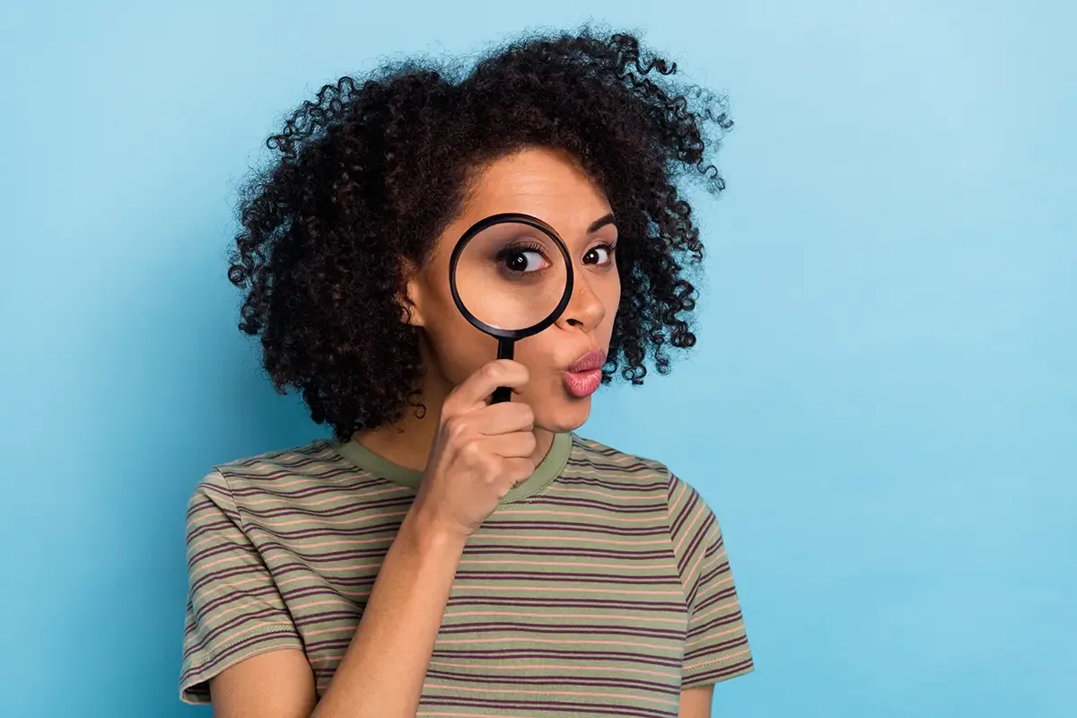 A woman with curly hair holds a magnifying glass up to her eye with a surprised, wide-eyed expression against a light blue background, conveying curiosity and close inspection.