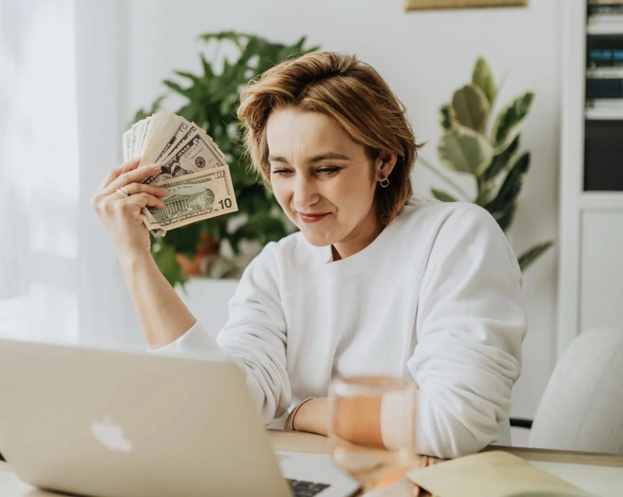 A woman with short hair, dressed in a white sweater, sits at a home office desk, holding a fan of U.S. dollar bills with a contemplative expression. She is looking at her MacBook, possibly reflecting on financial transactions or budgeting, in a room with a bright and airy decor highlighted by indoor plants.