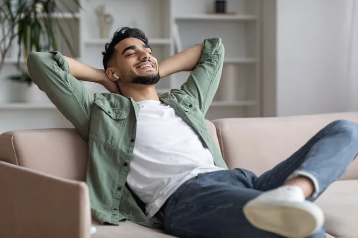 A man smiling while sitting on a couch with his hand behind his head and his legs crossed. He seems at peace.