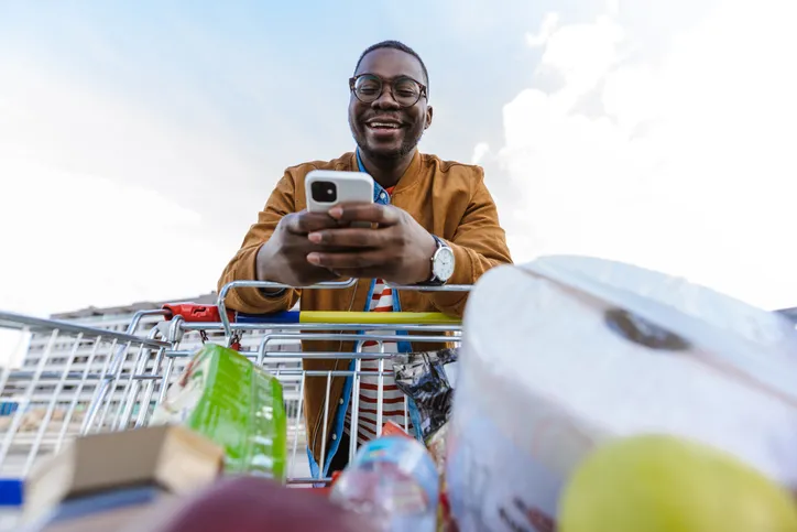 A person standing behind a shopping cart filled with groceries, using their smartphone and smiling. The photo is taken from a low angle, looking up at the person.