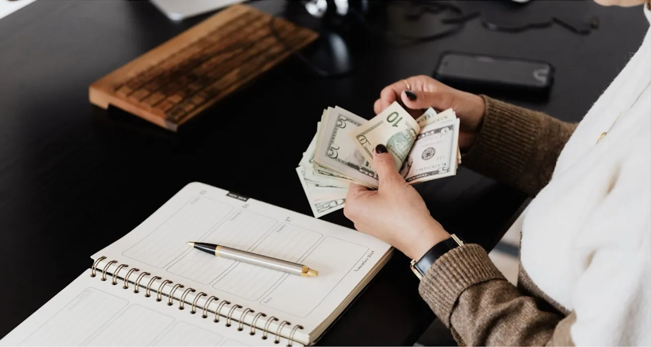 A person sits at a sleek black desk, counting a stack of U.S. dollar bills. Nearby, an open planner and a pen lay on the desk, suggesting a setting of financial planning or management. The person wears a casual beige sweater and a white blouse, indicating a professional yet relaxed environment.