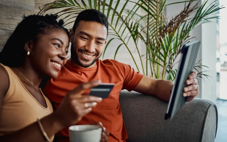 A couple with digital devices: An African American woman and a man of Asian descent are sitting together on a couch, looking at a tablet while the woman holds a credit card. Both are smiling and appear engaged, suggesting a pleasant online shopping or financial management session.