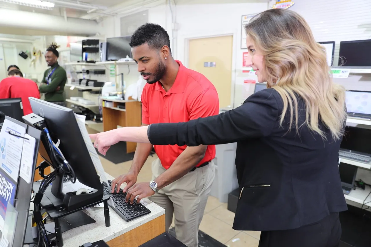 A district manager is pointing at a computer screen showing a Team Member what to do.