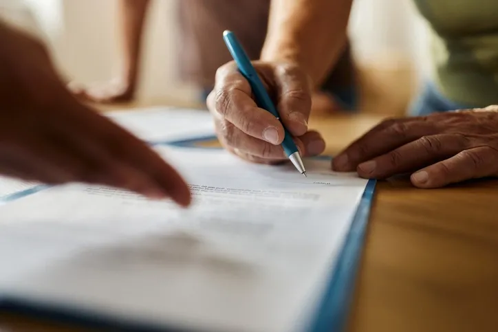 An image of a person signing documents: Close-up of hands, one holding a blue pen, signing or filling out a document. The focus on the paper and the hands conveys a sense of careful attention to important details.