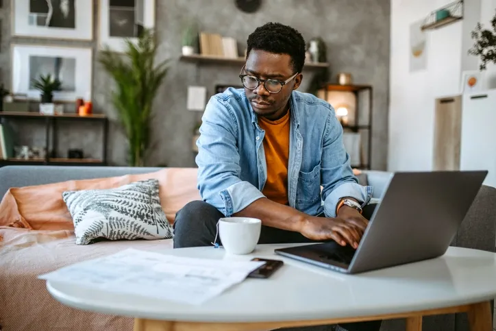 A man sitting in a living room, working on his laptop. He appears focused and is surrounded by a cozy home environment with a cup of coffee and some documents on the table.