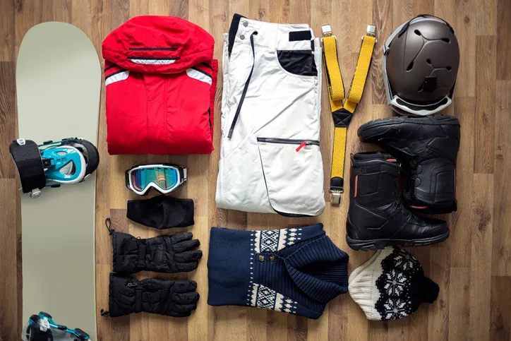 A variety of winter sports gear laid out on the floor, including a snowboard, boots, helmet, gloves, jacket, pants, goggles, and other accessories.