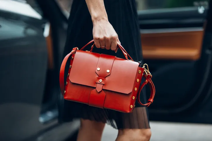 A women carrying a bright designer handbag from the waist down while stepping out of a car in a black dress.