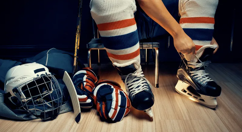 A hockey player putting on their ice skates in a locker room, with other hockey gear like gloves and a helmet nearby.