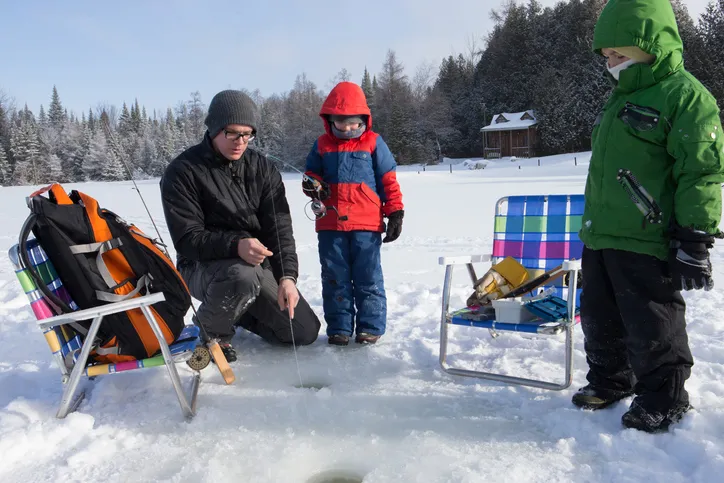 A father and his two children ice fishing. They are dressed warmly and standing near holes drilled in the ice.