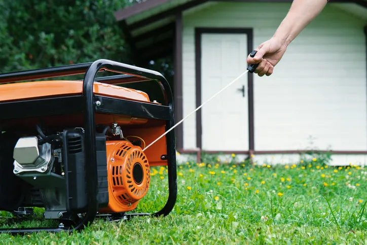 A person starting a portable generator by pulling the recoil starter cord. The generator is placed on grass, with a small building in the background.