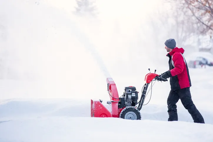 A person using a snowblower to clear a path through deep snow. The person is dressed in winter clothing and the snowblower is red.