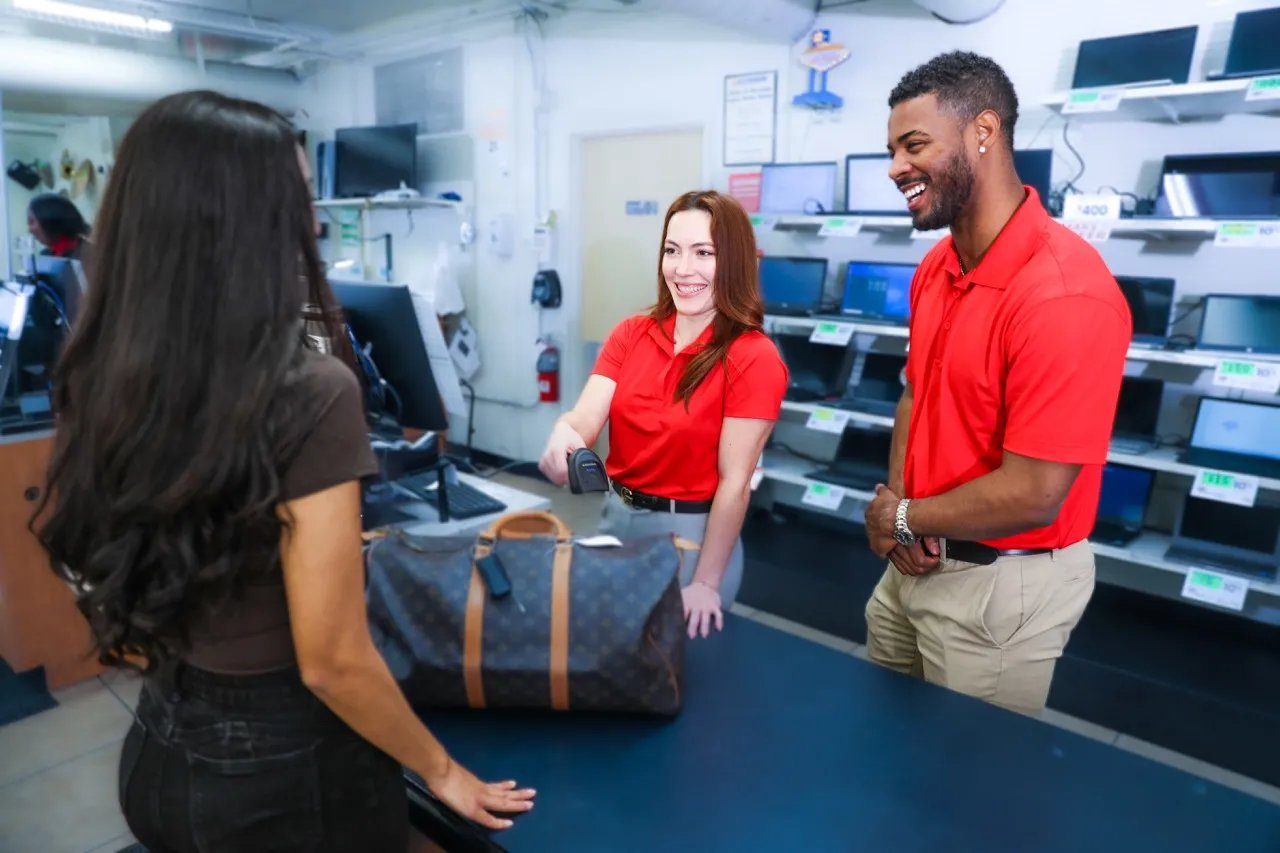 Two employees in red polo shirts smiling and assisting a customer with a designer handbag at a pawnshop counter.