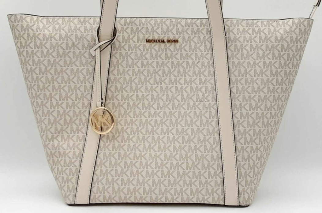 A large Michael Kors designer tote with a beige and white monogram pattern, featuring a circular logo charm and leather accents.