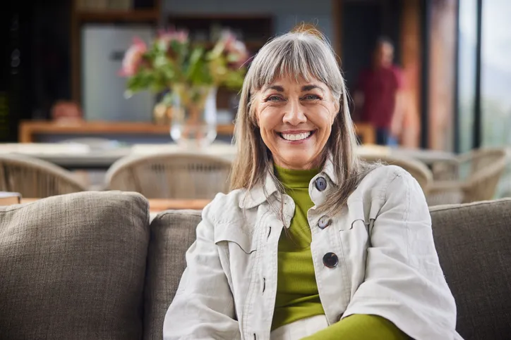 An older woman sitting on a couch, smiling warmly. The background suggests a comfortable home setting with some blurred elements, indicating a relaxed atmosphere.