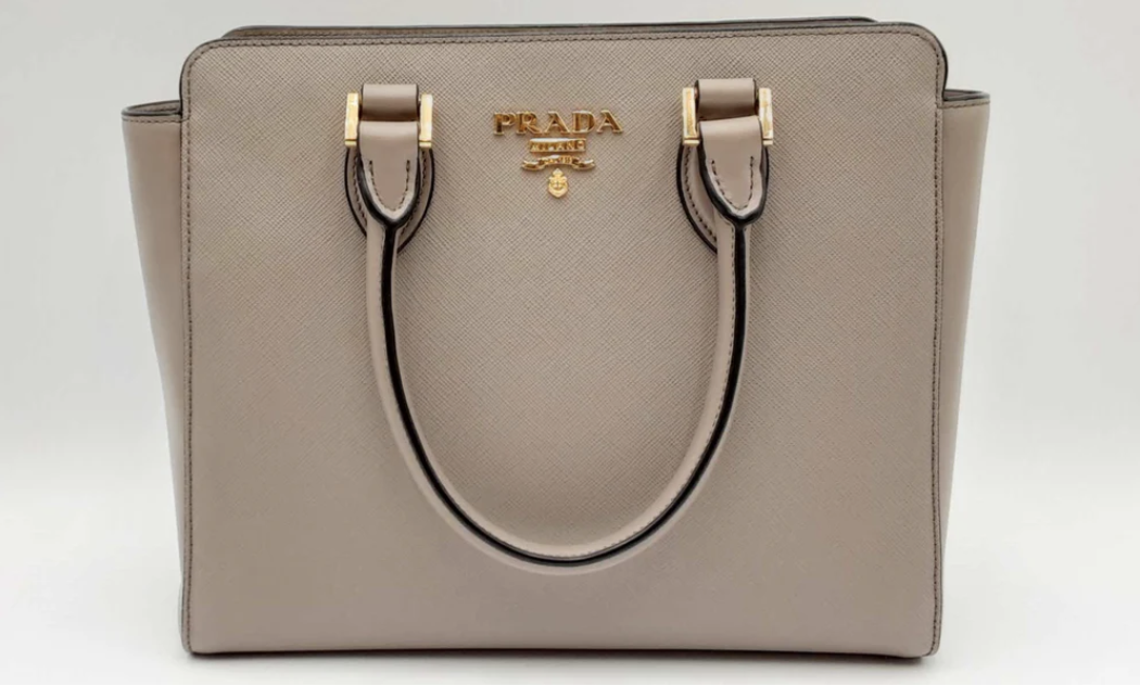 A gray Prada designer handbag with gold hardware, featuring dual handles and the brand logo in gold lettering.