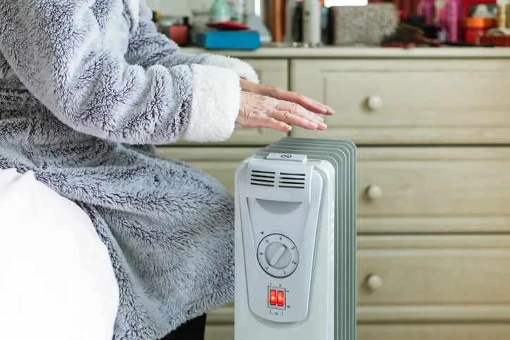 A person in a cozy robe warming their hands near a space heater, suggesting a cold indoor environment.