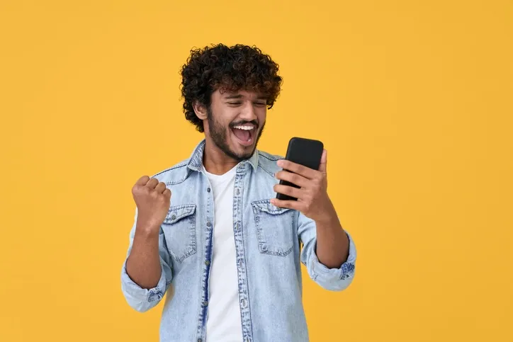A man celebrating with his phone: A young man with curly hair, wearing a light blue denim shirt, is exultantly cheering while looking at his smartphone, set against a bright yellow background. His jubilant expression and fist pump suggest he has received some very good news on his device.