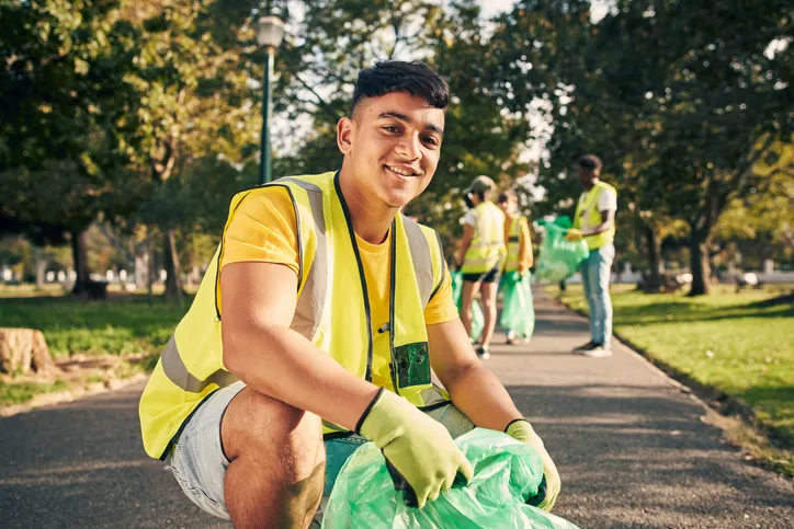 A young man wearing a high-visibility vest, smiling while participating in a park cleanup activity. Other volunteers can be seen in the background collecting trash.