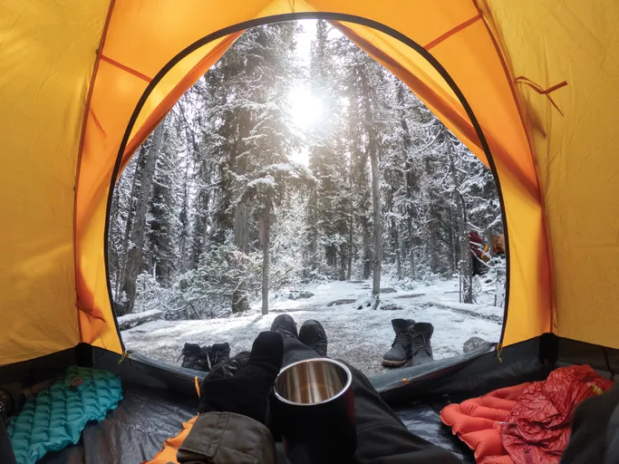 A view from inside a tent looking out at a snowy forest. The tent is yellow, and there are various camping gear items, including boots, visible inside the tent. A person is sitting inside the tent, holding a mug.