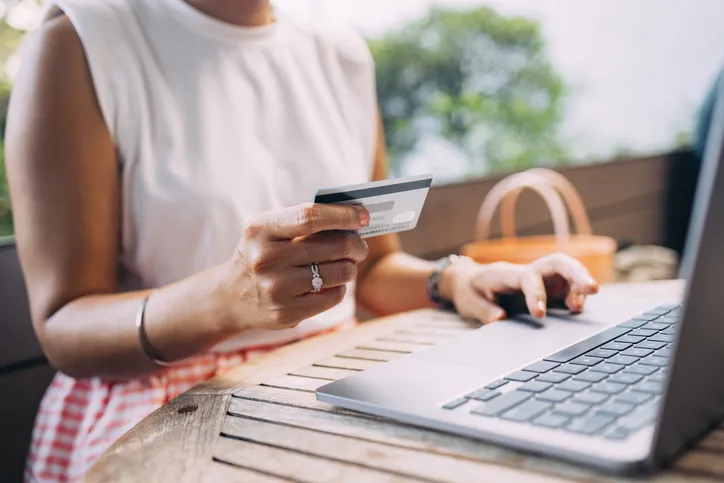 A close-up of a woman holding a credit card in one hand while typing on a laptop with the other. She is likely engaged in online shopping or making an online payment. The setting appears to be a casual outdoor environment.