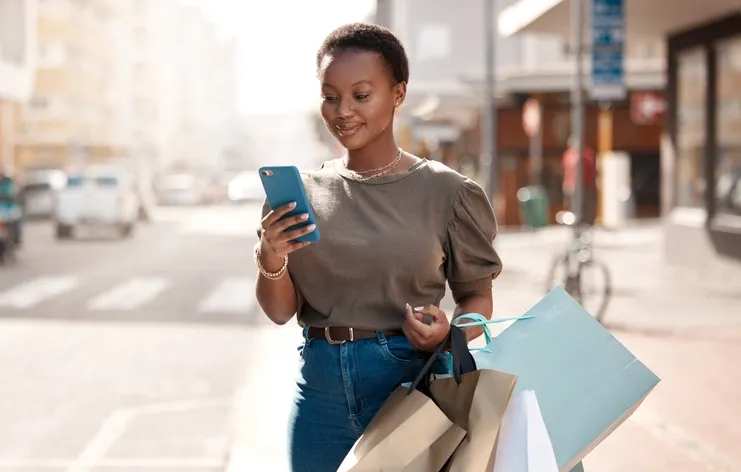 A woman standing outdoors, looking at her phone while holding several shopping bags. She seems to be enjoying a day out shopping, as suggested by her relaxed expression and the bright daylight setting.