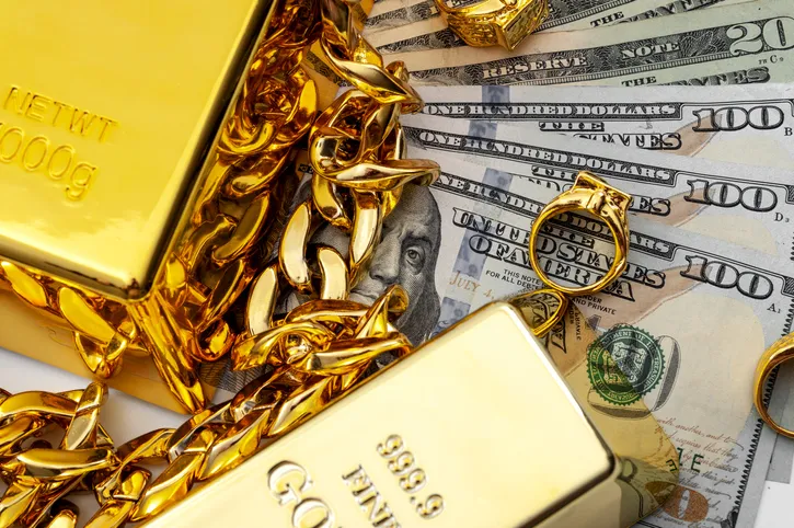 Gold bullion bars, gold chains, gold rings and a collection of $100 dollar bills are laid on a table.