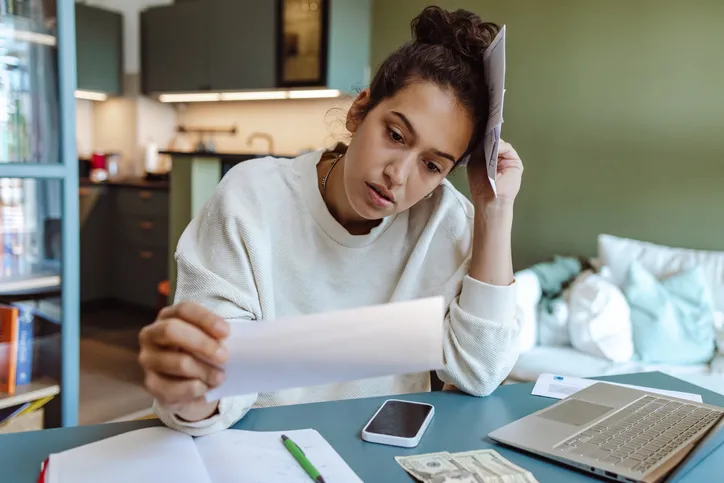 A women at a kitchen table is holding a piece of paper, likely a bill, while surrounded by papers, a phone, a laptop, a pen, and about sixty dollars of cash on the table. She is paying bills.