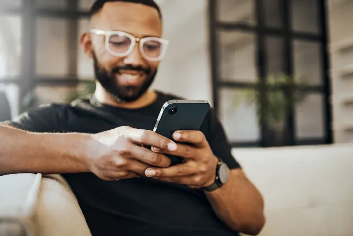 A man using a smartphone: A cheerful African American man wearing glasses and a black t-shirt is sitting comfortably on a couch, using a smartphone. His expression is content and focused, suggesting he's enjoying his interaction with the device.