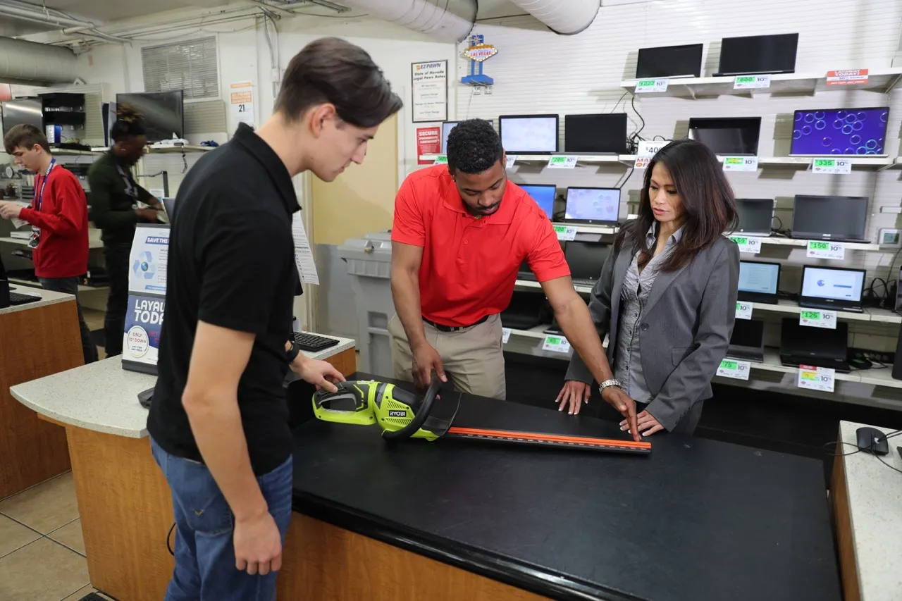 A customer is pawing a hedge trimmer while a Value Pawn Team Member examines it. A district manager is observing the transaction.