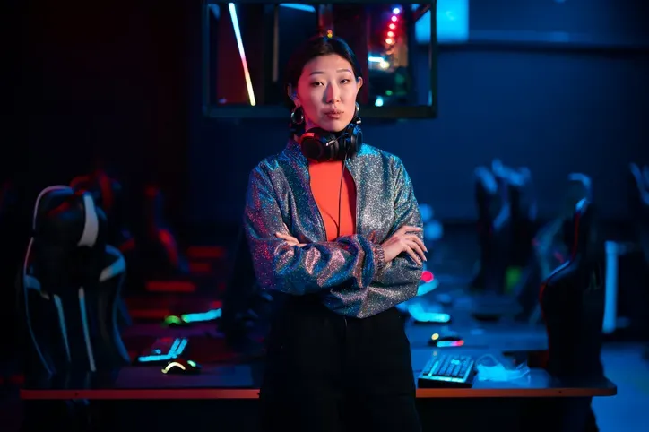 A woman standing in an esports gaming arena. She has headphones around her neck and is dressed in a stylish outfit, indicating her role as either a gamer or esports professional. The background shows multiple gaming stations.