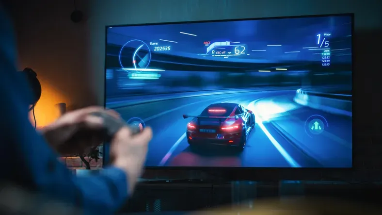 A person playing a racing video game. The focus is on the screen displaying a high-speed race, with the player's hands holding a controller in the foreground. The game interface includes speed and position indicators, typical of racing games.