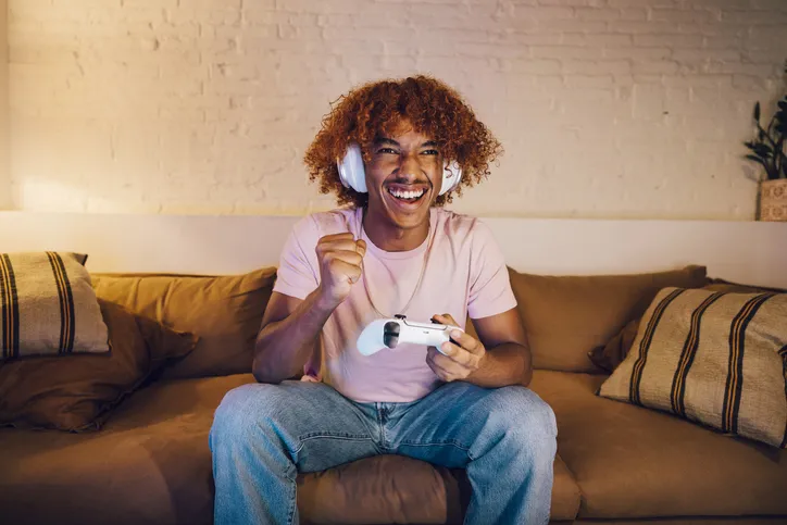 A young man with curly hair sitting on a couch, wearing headphones, and holding a video game controller while smiling and making a fist pump gesture. The background suggests a cozy and comfortable living space.