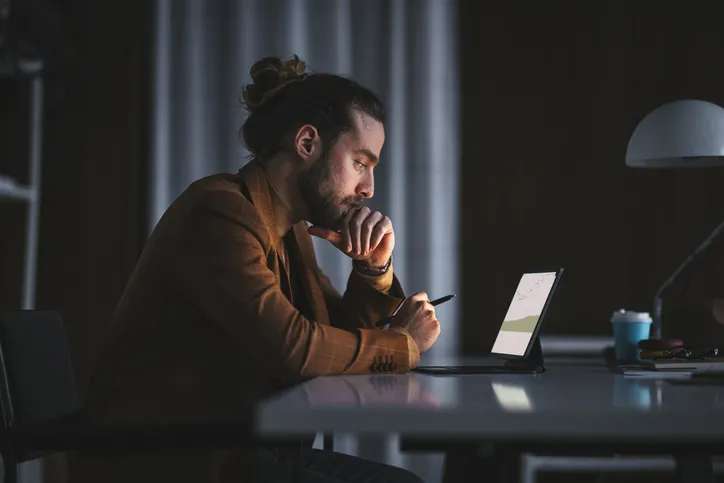 A man with a bun hairstyle concentrating deeply as he works on a tablet in a dimly lit office.