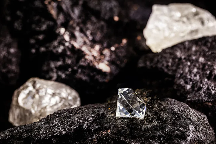 A raw diamond in its natural, rough state sitting atop a dark rock, with other uncut diamonds in the background.