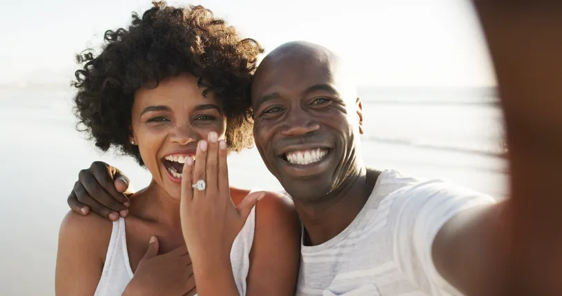 A couple taking a selfie on a beach, with the woman showing her engagement ring to the camera and smiling joyfully.