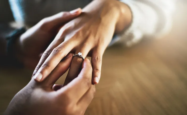 A man placing an engagement ring on a woman's finger. Her hands are in focus while their faces are blurred in the background.